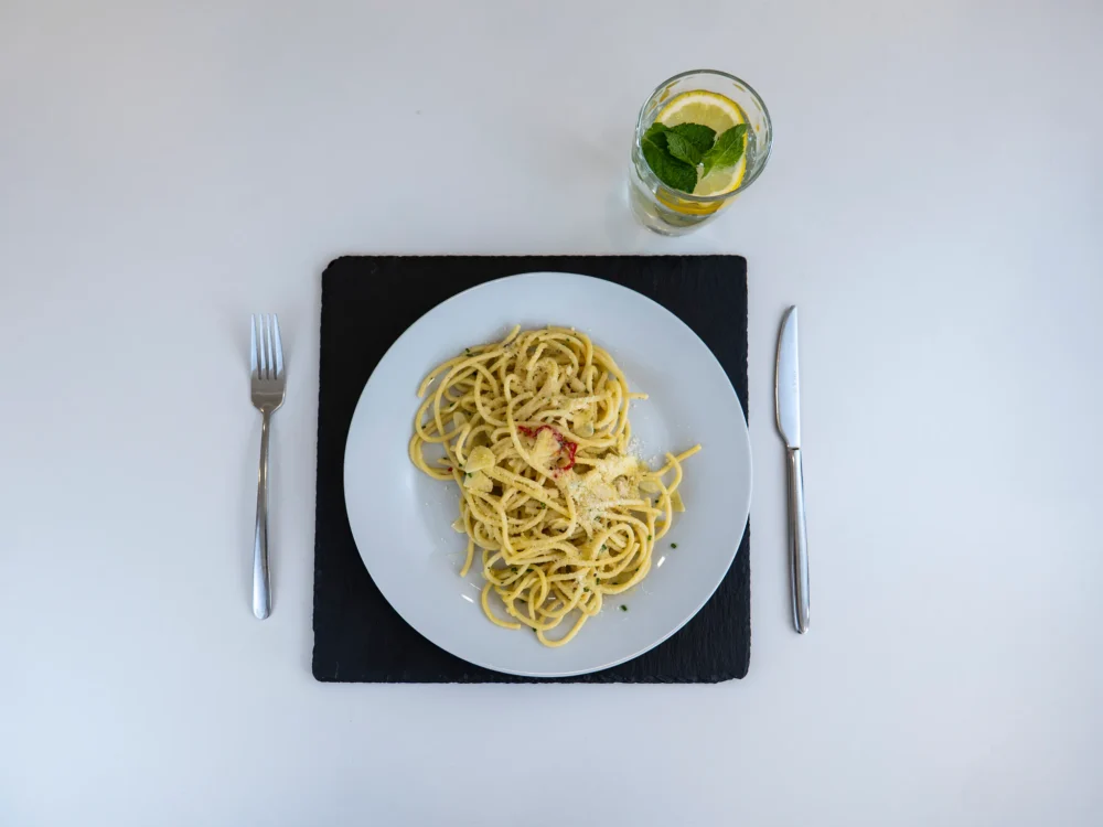 Špagety s chilli a parmazánem na bílém talíři, vedle vidlička, nůž a sklenice vody s citronem a mátou