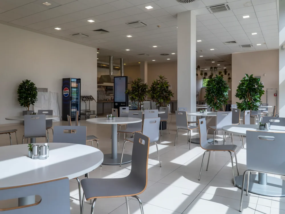 Jídelní prostor se slunečním světlem na podlaze, v pozadí lednice s nápoji, objednávkový kiosek a výdejní linka
