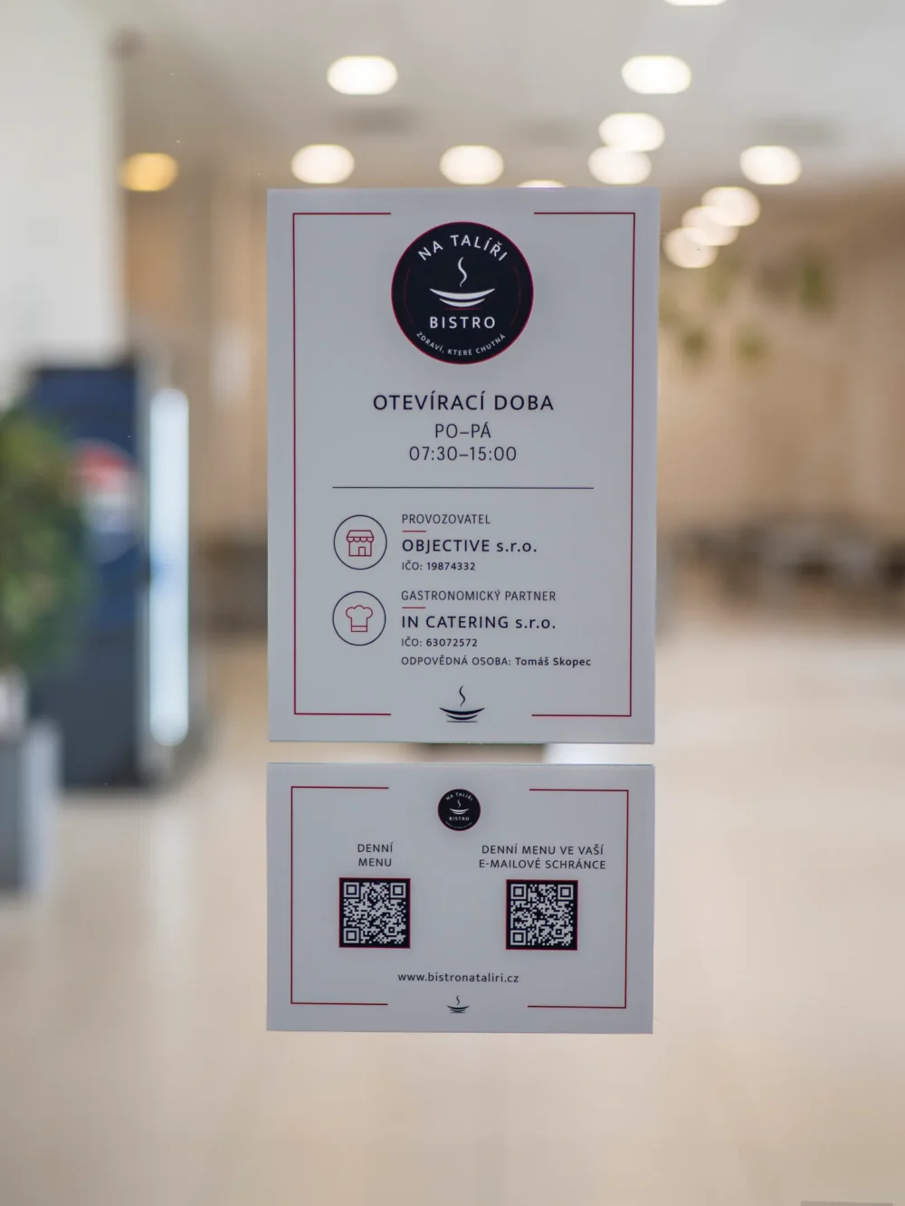Cedule na skleněných dveřích s otevírací dobou bistra, pondělí až pátek 7:30–15:00, a QR kódy na denní menu a jeho odběr e-mailem